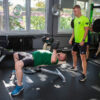 Trening z trenerem-personalnym a także trening z trenerem personalnym w parach w Gdańsku. Trener asystuje przy podnoszeniu przez mężczyznę hantli.